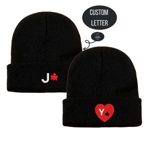 Peut inclure: Deux bonnets noirs avec des motifs brodés personnalisés. L'un présente la lettre "J" et une pièce de puzzle rouge, l'autre un cœur rouge avec la lettre "Y" et une pièce de puzzle rouge. Le texte "CUSTOM LETTER" est au-dessus des bonnets.