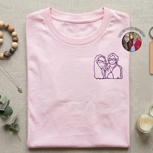 Può includere: Una t-shirt rosa chiaro con un ricamo a linee viola scuro di due persone sul petto sinistro. Un badge circolare nell'angolo in alto a destra mostra il testo CUSTOM PHOTO EMBROIDERY e una piccola fotografia di due persone.