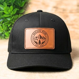 Peut inclure: Casquette de baseball noire avec un écusson en cuir marron. L'écusson présente un motif de montagne et d'arbres avec les mots "EXPLORE MORE" et "EST. 2024" en cercle. La casquette a une visière incurvée.