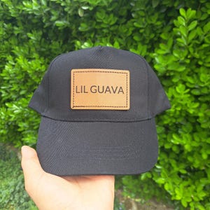 Può includere: Cappellino da baseball nero con visiera curva. Una toppa rettangolare con la scritta "LIL GUAVA" in maiuscolo è centrata sulla parte anteriore. La toppa è di colore marrone chiaro. Lo sfondo è un fogliame verde.