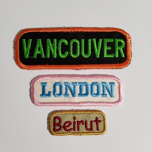 May include: Three rectangular embroidered patches. The top patch is black with green text reading VANCOUVER and an orange border. The middle patch is white with blue text reading LONDON and a pink border. The bottom patch is tan with red text reading Beirut and a gold border.