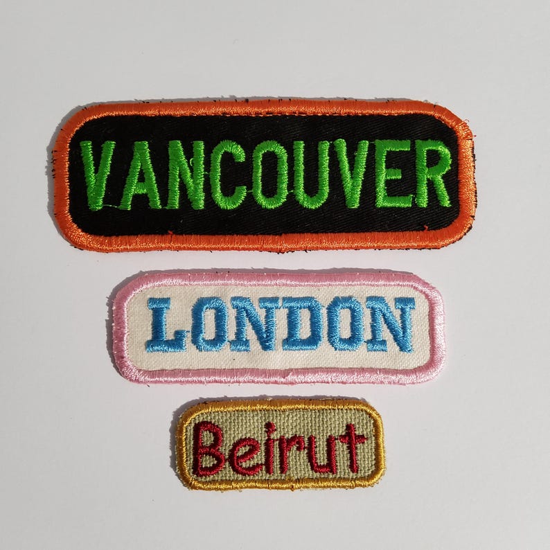 May include: Three rectangular embroidered patches. The top patch is black with green text reading VANCOUVER and an orange border. The middle patch is white with blue text reading LONDON and a pink border. The bottom patch is tan with red text reading Beirut and a gold border.