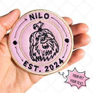 Peut inclure: Un écusson rond brodé rose avec le nom "NILO" en haut et "EST. 2024" en bas. Il présente une illustration en noir et blanc d'un chien. La bulle de texte dit "YOUR PET YOUR TEXT".
