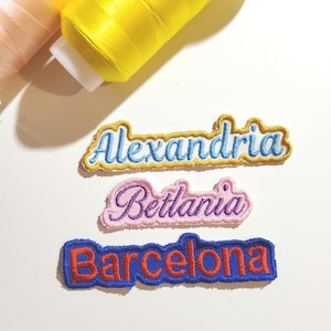 Puede incluir: Tres parches bordados con los nombres Alexandria en azul claro con borde dorado, Betlania en morado con borde rosa y Barcelona en rojo con borde azul oscuro, situados debajo de dos carretes de hilo de color amarillo y melocotón.