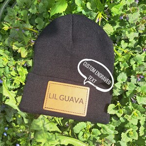 Può includere: Berretto nero con una toppa in pelle marrone con la scritta "LIL GUAVA". Un adesivo a forma di fumetto bianco con la scritta "CUSTOM ENGRAVED TEXT" è sul berretto. Il berretto è su foglie verdi.