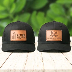 Peut inclure: Deux casquettes de baseball noires avec des écussons en cuir marron. L'un présente un contour de bâtiment et le texte "AETHEL CONSTRUCTION EST. 1998." L'autre affiche une fourchette et un couteau avec "OLIVE BRANCH BISTRO CROATIA."