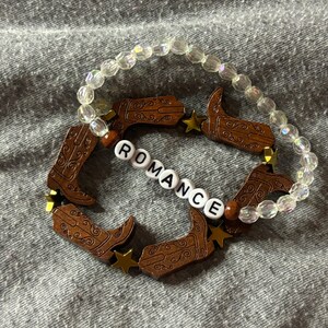 May include: A white beaded bracelet with brown cowboy boot charms and gold star charms. The bracelet also has white letter beads that spell out the word "ROMANCE".