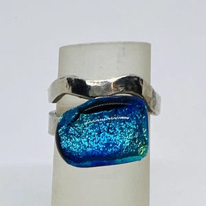 May include: A silver ring with a dichroic glass stone. The stone is a vibrant blue with hints of green and a textured surface. The ring band has a hammered texture.