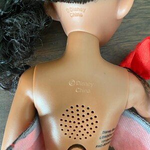 以下が含まれることがあります： 黒髪の肌色の人形。人形の背面が見えます。人形にはスピーカーと電池収納部があります。「Disney China」の文字が人形の背中に印刷されています。