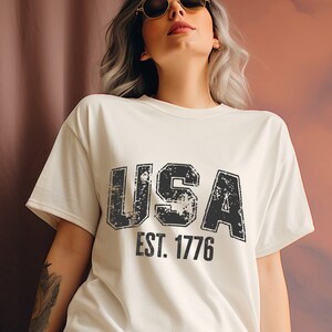 White t-shirt with a black distressed graphic that says 'USA EST. 1776'.