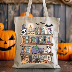Puede incluir: Bolso tote de lona beige con un dise&ntilde;o de estanter&iacute;a de Halloween. La ilustraci&oacute;n incluye una calavera con sombrero de bruja, libros, un caldero, un fantasma y una telara&ntilde;a. El bolso tiene un asa de color claro y cuelga sobre un fondo de madera.
