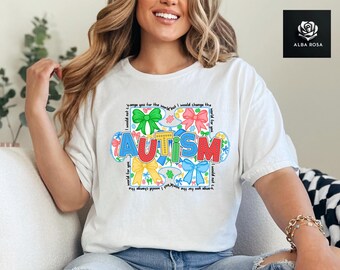 May include: A white short-sleeved t-shirt featuring a colorful graphic of puzzle pieces and bows. The word AUTISM is printed in large, multi-colored letters. The text I would not change you for the world but I would change the world for you is written in a square border around the central design.