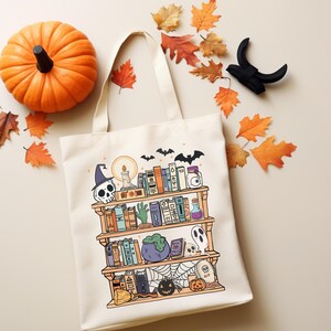 Puede incluir: Un bolso tote crema con una ilustraci&oacute;n de estanter&iacute;a con tem&aacute;tica de Halloween. El dise&ntilde;o incluye una calavera, libros, una botella de poci&oacute;n, un fantasma y una calabaza. Una calabaza naranja y hojas de oto&ntilde;o se colocan alrededor de la bolsa.