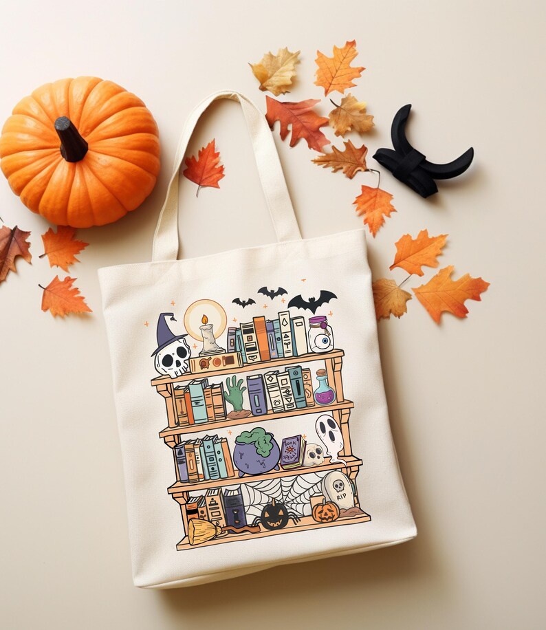 Puede incluir: Un bolso tote crema con una ilustraci&oacute;n de estanter&iacute;a con tem&aacute;tica de Halloween. El dise&ntilde;o incluye una calavera, libros, una botella de poci&oacute;n, un fantasma y una calabaza. Una calabaza naranja y hojas de oto&ntilde;o se colocan alrededor de la bolsa.