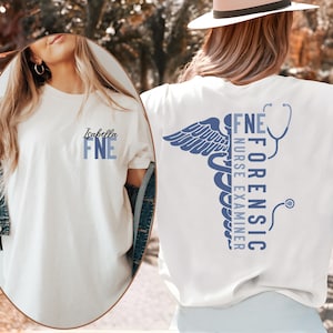 May include: A white t-shirt featuring a blue graphic design. The front displays the name Isabella above the letters FNE. The back features a large caduceus symbol, a stethoscope, and the vertical text Forensic Nurse Examiner in blue.