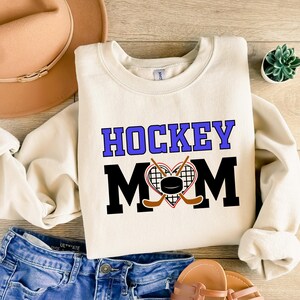 Puede incluir: Una sudadera de color crema con el texto "HOCKEY MOM" en letras negras y moradas. Un coraz&oacute;n con palos de hockey y un disco est&aacute; en el centro del texto.
