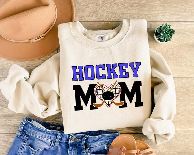 Puede incluir: Una sudadera de color crema con el texto "HOCKEY MOM" en letras negras y moradas. Un coraz&oacute;n con palos de hockey y un disco est&aacute; en el centro del texto.
