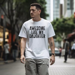May include: White t-shirt with the text "AWESOME LIKE MY DAUGHTER" in a distressed black font. The shirt is worn by a person walking down a city street. The person is wearing gray cargo pants and a watch.