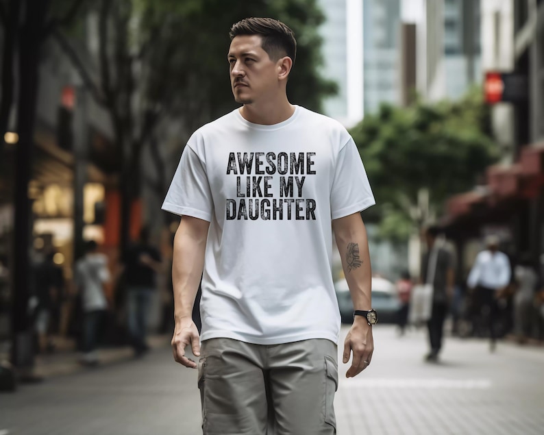 May include: White t-shirt with the text "AWESOME LIKE MY DAUGHTER" in a distressed black font. The shirt is worn by a person walking down a city street. The person is wearing gray cargo pants and a watch.