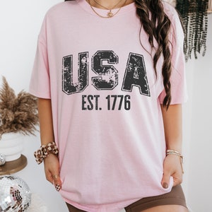 May include: A pink t-shirt with a distressed black graphic that says "USA EST. 1776".