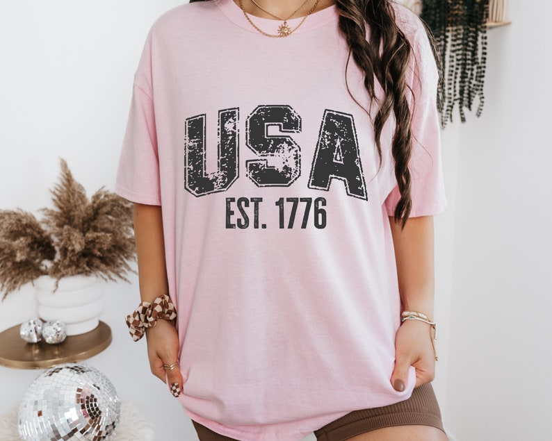 May include: A pink t-shirt with a distressed black graphic that says "USA EST. 1776".