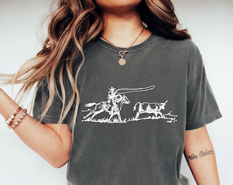 May include: A charcoal gray short-sleeve t-shirt featuring a white line art graphic of a cowboy on horseback roping a steer. The design depicts the rider in a hat holding a lasso, with the animal running ahead, all set against a plain background.