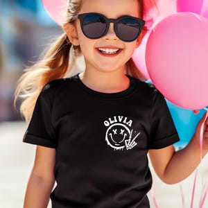 May include: A black t-shirt with a white graphic of a smiley face with an X for eyes and a skeleton hand making a peace sign. The text "OLIVIA" is above the graphic.