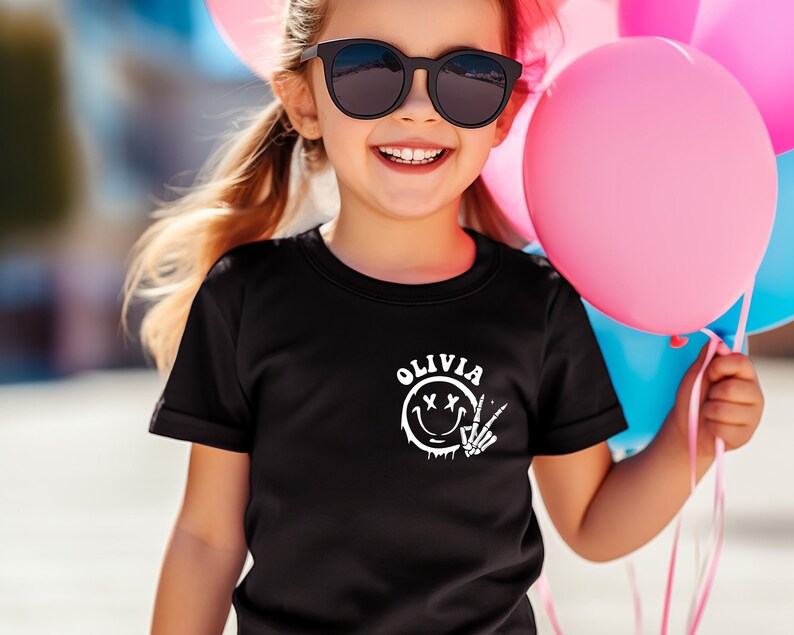 May include: A black t-shirt with a white graphic of a smiley face with an X for eyes and a skeleton hand making a peace sign. The text "OLIVIA" is above the graphic.
