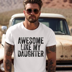May include: White t-shirt with the text "AWESOME LIKE MY DAUGHTER" in black, distressed lettering. The shirt is worn by a person with tattoos, sunglasses, and a beard. A vintage truck is in the background.