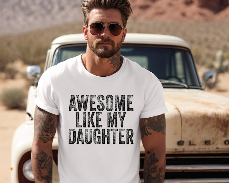 May include: White t-shirt with the text "AWESOME LIKE MY DAUGHTER" in black, distressed lettering. The shirt is worn by a person with tattoos, sunglasses, and a beard. A vintage truck is in the background.