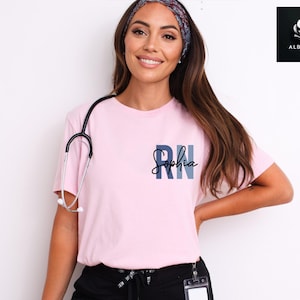 May include: A pink t-shirt with a blue and gray "RN" design and the name "Sophia" written in cursive. The shirt is being worn by a person with long brown hair and a headband.