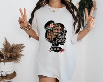 May include: A white short-sleeve t-shirt featuring a black silhouette of a head filled with a bookshelf graphic. The text reads: & Into the library she goes to lose her MIND and find her SOUL. Colorful floral accents surround the silhouette.