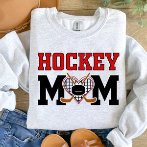 Puede incluir: Sudadera gris con el texto "HOCKEY MOM" en letras rojas y negras. Un coraz&oacute;n con un disco de hockey y palos de hockey cruzados se encuentra en el centro del texto.