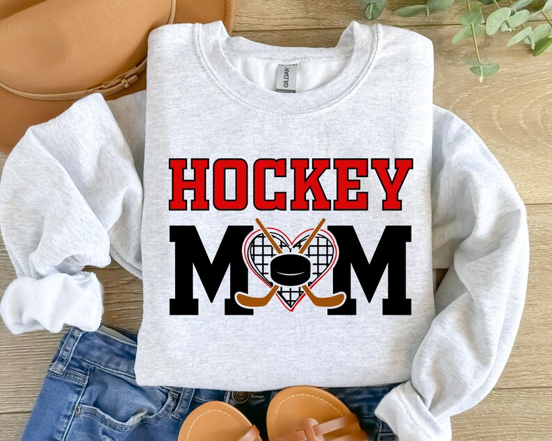 Puede incluir: Sudadera gris con el texto "HOCKEY MOM" en letras rojas y negras. Un coraz&oacute;n con un disco de hockey y palos de hockey cruzados se encuentra en el centro del texto.