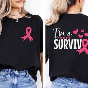 Op de afbeelding: Zwart T-shirt met een roze lint op de voorkant en de tekst "I'm a SURVIVOR" met een roze lint op de achterkant. Het shirt is een symbool voor bewustwording van borstkanker.