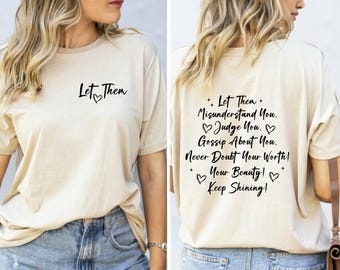 May include: A beige t-shirt featuring black text and heart graphics. The front displays the words Let Them with a heart. The back features the text Let Them Misunderstand You. Judge You. Gossip About You. Never Doubt Your Worth! Your Beauty! Keep Shining!