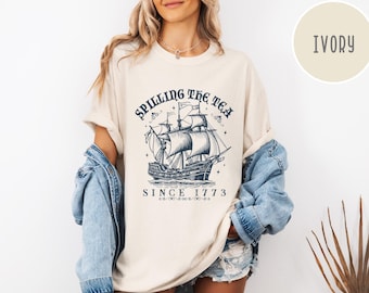 May include: An ivory t-shirt featuring a dark blue graphic of a sailing ship with the text "SPILLING THE TEA" arched above and "SINCE 1773" below. The design includes small star accents and decorative flourishes surrounding the detailed ship illustration.
