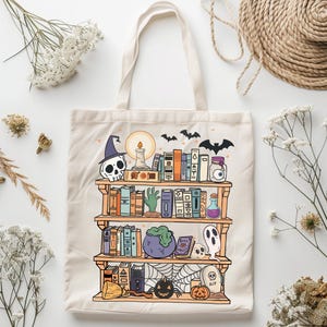 A white canvas tote bag with a printed design of a bookshelf filled with books, a cauldron, a skull, a ghost, a spiderweb, a pumpkin, and a tombstone with the word 'RIP' on it. The bookshelf is decorated with bats, a candle, and a potion bottle with an eyeball inside. The design is in a cartoon style with bright colors.
