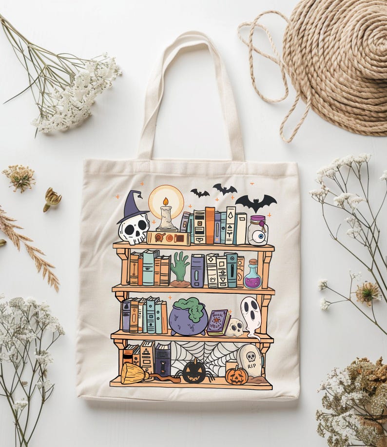 A white canvas tote bag with a printed design of a bookshelf filled with books, a cauldron, a skull, a ghost, a spiderweb, a pumpkin, and a tombstone with the word 'RIP' on it. The bookshelf is decorated with bats, a candle, and a potion bottle with an eyeball inside. The design is in a cartoon style with bright colors.