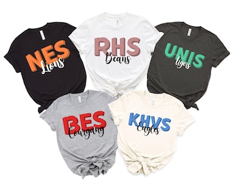 May include: Five short-sleeve t-shirts in black, white, dark gray, heather gray, and cream. Each shirt features bold block letters and cursive text: NES Lions, RHS Beans, UNIS Tigers, BES Courgang, and KHVS Eagles in orange, pink, green, red, and blue.