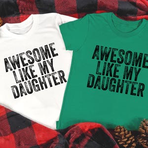 May include: Two short-sleeved t-shirts, one white and one green, with the text "AWESOME LIKE MY DAUGHTER" printed in black. The shirts are displayed on a red and black plaid blanket, with pine cones.
