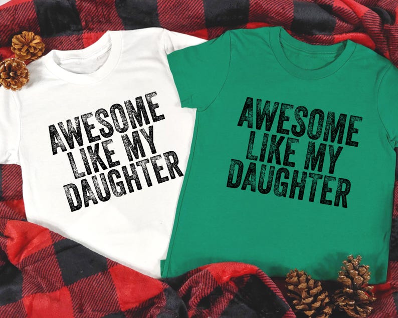 May include: Two short-sleeved t-shirts, one white and one green, with the text "AWESOME LIKE MY DAUGHTER" printed in black. The shirts are displayed on a red and black plaid blanket, with pine cones.