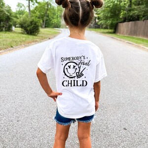 May include: White t-shirt with black text that reads "Somebody's Feral Child" and a black smiley face with an X for eyes and a peace sign.