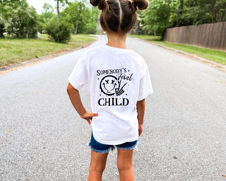 May include: White t-shirt with black text that reads "Somebody's Feral Child" and a black smiley face with an X for eyes and a peace sign.