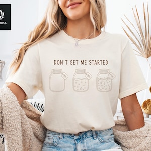 May include: Cream-colored t-shirt with the text "DON'T GET ME STARTED" above three jar illustrations. The jars have varying contents and tags. The shirt is a casual, everyday wear item.