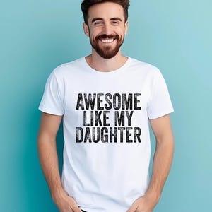 May include: White t-shirt with the text "AWESOME LIKE MY DAUGHTER" in a bold, distressed black font. The shirt is worn by a person with a beard, smiling against a light blue background. A casual, everyday wear.