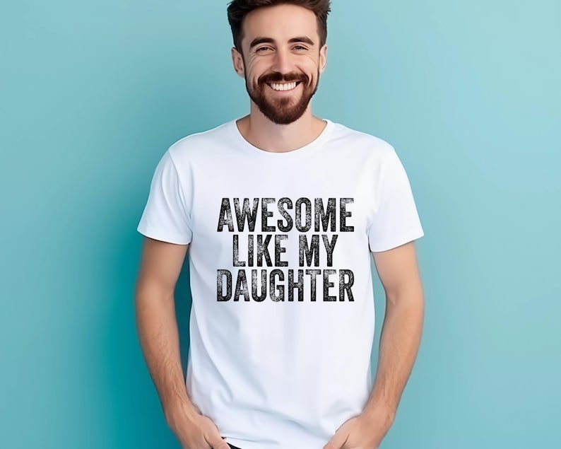 May include: White t-shirt with the text "AWESOME LIKE MY DAUGHTER" in a bold, distressed black font. The shirt is worn by a person with a beard, smiling against a light blue background. A casual, everyday wear.