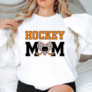 Puede incluir: Sudadera blanca con el texto "HOCKEY MOM" en letras negras y naranjas. Un coraz&oacute;n con palos de hockey y un disco est&aacute; en el centro del texto.