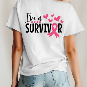 Op de afbeelding: Wit T-shirt met de tekst "I'm a SURVIVOR" in zwart met een roze lint en roze harten.
