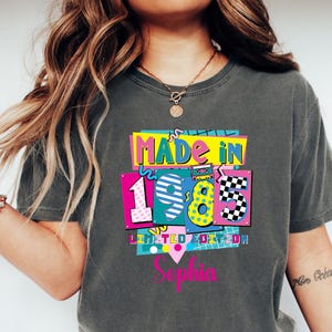 May include: Dark gray t-shirt with a colorful graphic that reads "MADE IN 1985 LIMITED EDITION" with the name "Sophia" below. The design features vibrant colors like pink, yellow, blue, and black, creating a retro aesthetic.